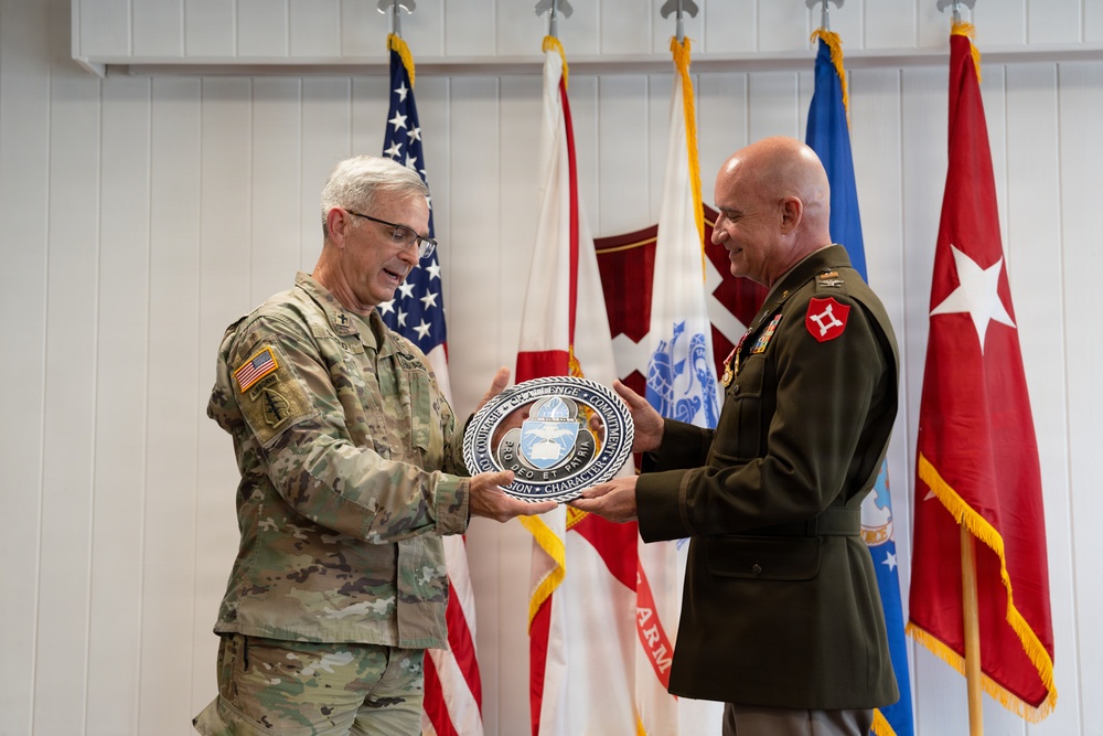 Two senior U.S. Army officers are engaged in a formal presentation ceremony, with one officer in MultiCam/OCP combat uniform and the other in Army Green Service uniform. They are jointly holding a large commemorative medallion or plaque featuring text including 'Courage,' 'Challenge,' 'Commitment,' 'Character,' and 'Pro Deo Et Patri.' Multiple military flags and unit colors are displayed in the background.