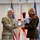 Col. Finch Concludes Distinguished Career, Leaves Lasting Legacy as Command State Chaplain Two senior U.S. Army officers are engaged in a formal presentation ceremony, with one officer in MultiCam/OCP combat uniform and the other in Army Green Service uniform. They are jointly holding a large commemorative medallion or plaque featuring text including 'Courage,' 'Challenge,' 'Commitment,' 'Character,' and 'Pro Deo Et Patri.' Multiple military flags and unit colors are displayed in the background.
