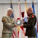 Two senior U.S. Army officers are engaged in a formal presentation ceremony, with one officer in MultiCam/OCP combat uniform and the other in Army Green Service uniform. They are jointly holding a large commemorative medallion or plaque featuring text including 'Courage,' 'Challenge,' 'Commitment,' 'Character,' and 'Pro Deo Et Patri.' Multiple military flags and unit colors are displayed in the background.