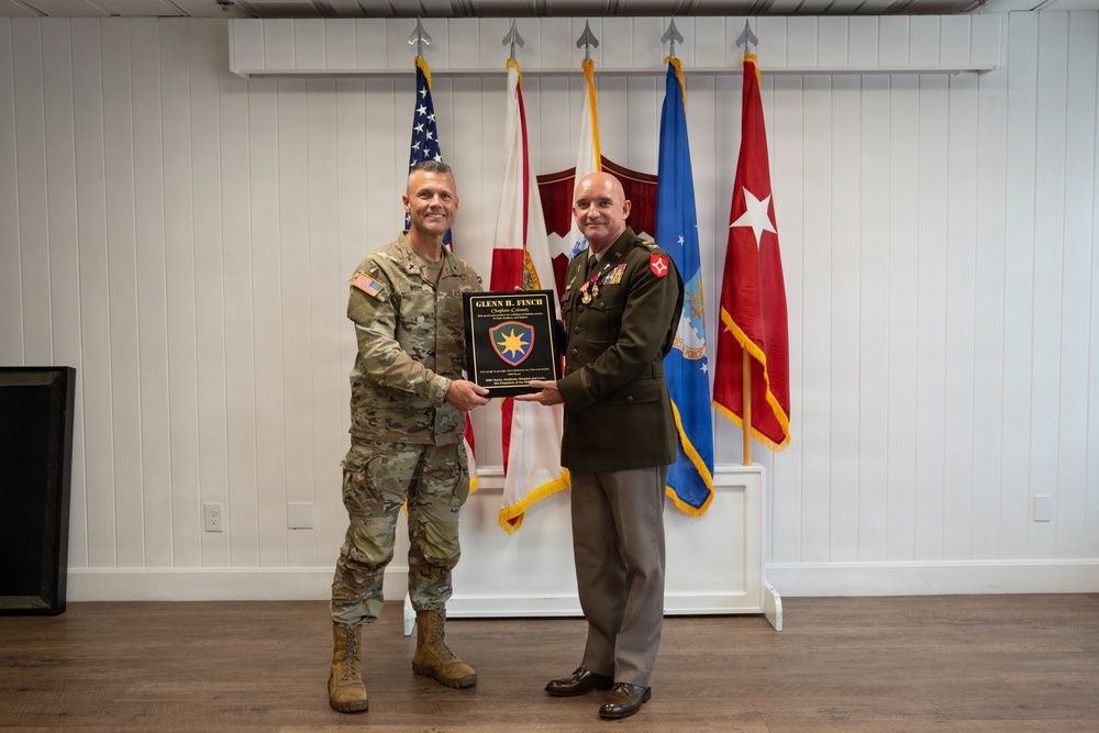 Two military personnel are posing together while jointly holding a commemorative plaque inscribed with 'Glenn H. Finch, Chaplain Colonel.' The individual on the left is wearing U.S. Army combat uniform (OCP/multicam), while the individual on the right is wearing a U.S. Marine Corps dress green uniform with medals and a general officer's rank insignia. Several military flags are displayed in the background.