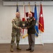 Two military personnel are posing together while jointly holding a commemorative plaque inscribed with 'Glenn H. Finch, Chaplain Colonel.' The individual on the left is wearing U.S. Army combat uniform (OCP/multicam), while the individual on the right is wearing a U.S. Marine Corps dress green uniform with medals and a general officer's rank insignia. Several military flags are displayed in the background.