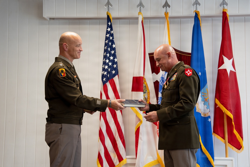 Two U.S. Army officers in olive green dress uniforms are exchanging or presenting what appears to be a framed certificate or award during a formal military ceremony. The setting includes multiple flags including the U.S. flag, state flags, and military unit flags displayed behind them. The officer on the right is wearing multiple service medals and ribbons, suggesting this may be a retirement, promotion, or commendation ceremony.