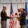 Col. Finch Concludes Distinguished Career, Leaves Lasting Legacy as Command State Chaplain Two U.S. Army officers in olive green dress uniforms are exchanging or presenting what appears to be a framed certificate or award during a formal military ceremony. The setting includes multiple flags including the U.S. flag, state flags, and military unit flags displayed behind them. The officer on the right is wearing multiple service medals and ribbons, suggesting this may be a retirement, promotion, or commendation ceremony.