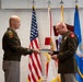 Two U.S. Army officers in olive green dress uniforms are exchanging or presenting what appears to be a framed certificate or award during a formal military ceremony. The setting includes multiple flags including the U.S. flag, state flags, and military unit flags displayed behind them. The officer on the right is wearing multiple service medals and ribbons, suggesting this may be a retirement, promotion, or commendation ceremony.