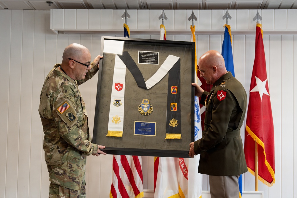 Two military officers are holding a framed shadow box display during what appears to be a formal military ceremony. The shadow box contains military insignia, patches, a stole/sash, and commemorative plaques, likely presented as a retirement or farewell gift. The setting includes multiple military flags in the background.