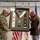 Col. Finch Concludes Distinguished Career, Leaves Lasting Legacy as Command State Chaplain Two military officers are holding a framed shadow box display during what appears to be a formal military ceremony. The shadow box contains military insignia, patches, a stole/sash, and commemorative plaques, likely presented as a retirement or farewell gift. The setting includes multiple military flags in the background.