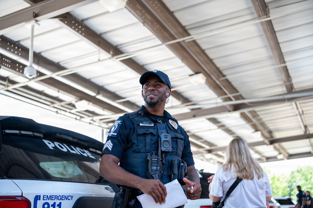 A police sergeant in full uniform stands near a marked police vehicle under a covered structure, holding papers and appearing to look upward with a relaxed expression. A body camera is visible on his chest, and he is wearing an earpiece. A civilian with blonde hair is visible in the background to the right.