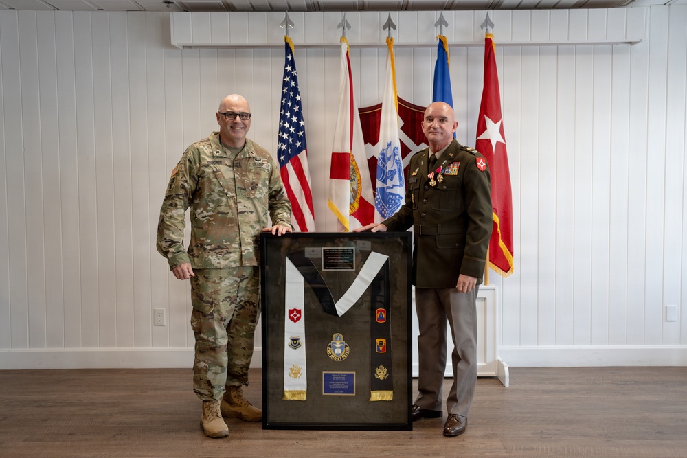 Two U.S. military personnel pose together holding a framed shadow box display containing military insignia, patches, a sash, and commemorative plaques. The individual on the left wears Army Combat Uniform (ACU/OCP multicam), while the individual on the right wears a Class A dress uniform with multiple medals and ribbons. Several military and U.S. flags are displayed in the background, suggesting a formal ceremony or retirement event.
