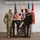 Col. Finch Concludes Distinguished Career, Leaves Lasting Legacy as Command State Chaplain Two U.S. military personnel pose together holding a framed shadow box display containing military insignia, patches, a sash, and commemorative plaques. The individual on the left wears Army Combat Uniform (ACU/OCP multicam), while the individual on the right wears a Class A dress uniform with multiple medals and ribbons. Several military and U.S. flags are displayed in the background, suggesting a formal ceremony or retirement event.