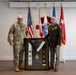 Two U.S. military personnel pose together holding a framed shadow box display containing military insignia, patches, a sash, and commemorative plaques. The individual on the left wears Army Combat Uniform (ACU/OCP multicam), while the individual on the right wears a Class A dress uniform with multiple medals and ribbons. Several military and U.S. flags are displayed in the background, suggesting a formal ceremony or retirement event.