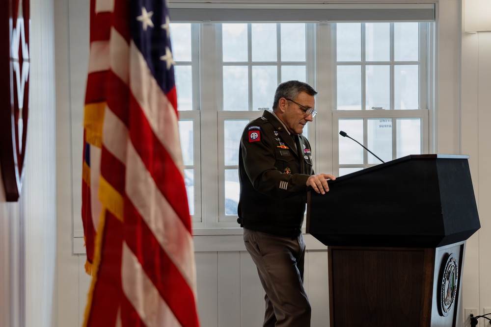 A U.S. Army officer in dress uniform stands at a dark podium, appearing to review notes or prepare to speak. The setting appears to be an official military ceremony or briefing held indoors. An American flag is prominently displayed to the left of the frame.