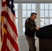 A U.S. Army officer in dress uniform stands at a dark podium, appearing to review notes or prepare to speak. The setting appears to be an official military ceremony or briefing held indoors. An American flag is prominently displayed to the left of the frame.