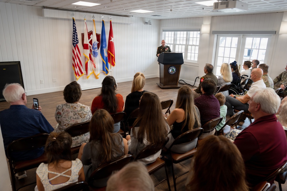 A military officer in uniform is delivering a speech at a podium during what appears to be an official military ceremony or promotion event. An audience of approximately 20-25 civilians and military personnel are seated in folding chairs facing the speaker. Multiple military flags are displayed prominently behind the podium against a white paneled wall.