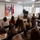 Col. Finch Concludes Distinguished Career, Leaves Lasting Legacy as Command State Chaplain A military officer in uniform is delivering a speech at a podium during what appears to be an official military ceremony or promotion event. An audience of approximately 20-25 civilians and military personnel are seated in folding chairs facing the speaker. Multiple military flags are displayed prominently behind the podium against a white paneled wall.
