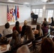 A military officer in uniform is delivering a speech at a podium during what appears to be an official military ceremony or promotion event. An audience of approximately 20-25 civilians and military personnel are seated in folding chairs facing the speaker. Multiple military flags are displayed prominently behind the podium against a white paneled wall.