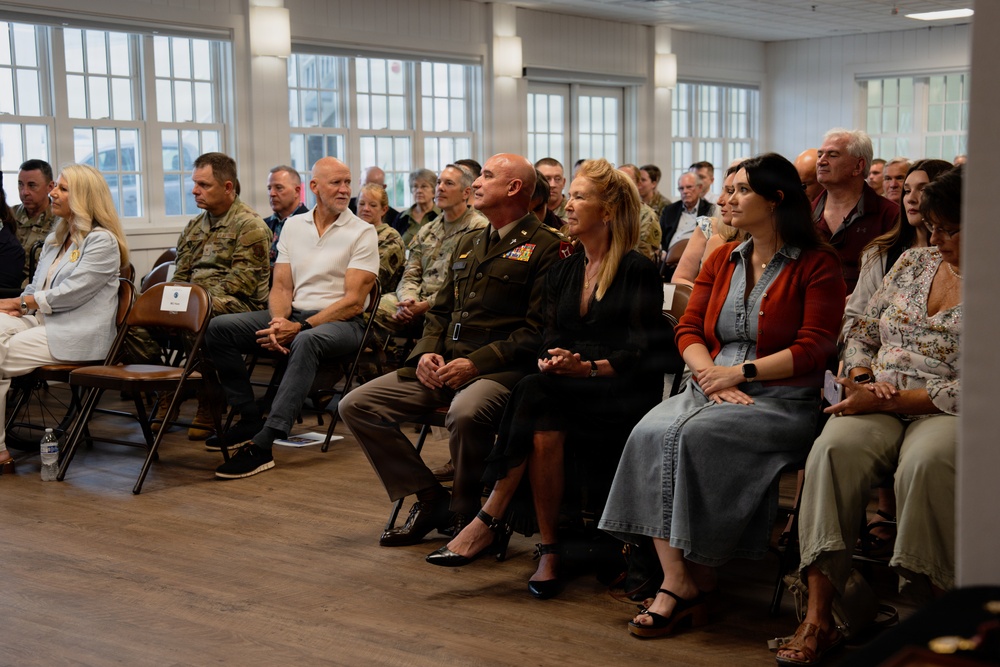 A formal indoor gathering featuring a mix of military personnel in uniform and civilians seated in rows of folding chairs. The audience appears to be attentively watching a presentation or ceremony taking place off-camera. The setting is a well-lit room with large windows, suggesting a community hall or event venue.