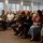 Col. Finch Concludes Distinguished Career, Leaves Lasting Legacy as Command State Chaplain A formal indoor gathering featuring a mix of military personnel in uniform and civilians seated in rows of folding chairs. The audience appears to be attentively watching a presentation or ceremony taking place off-camera. The setting is a well-lit room with large windows, suggesting a community hall or event venue.