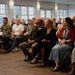 A formal indoor gathering featuring a mix of military personnel in uniform and civilians seated in rows of folding chairs. The audience appears to be attentively watching a presentation or ceremony taking place off-camera. The setting is a well-lit room with large windows, suggesting a community hall or event venue.