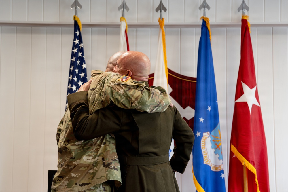 Two military personnel in uniform are embracing in what appears to be an official ceremony. They are standing in front of a display of military flags including the U.S. flag, an Air Force flag, and a general officer flag. The setting suggests a formal military event such as a change of command, retirement, or promotion ceremony.