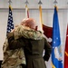 Two military personnel in uniform are embracing in what appears to be an official ceremony. They are standing in front of a display of military flags including the U.S. flag, an Air Force flag, and a general officer flag. The setting suggests a formal military event such as a change of command, retirement, or promotion ceremony.