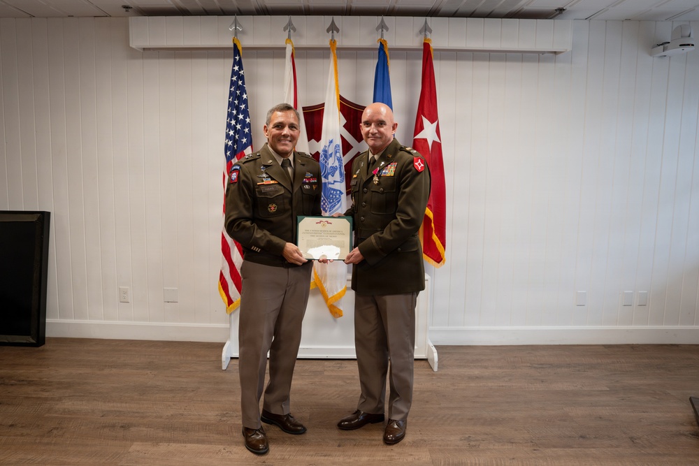 Two U.S. Army officers in service dress uniforms are posing together while jointly holding a certificate or award document. The setting appears to be a formal military ceremony room with multiple flags displayed in the background. This appears to be an awards or recognition ceremony.