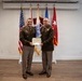 Two U.S. Army officers in service dress uniforms are posing together while jointly holding a certificate or award document. The setting appears to be a formal military ceremony room with multiple flags displayed in the background. This appears to be an awards or recognition ceremony.