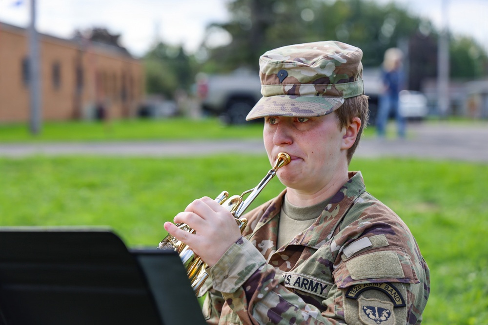 A U.S. Army soldier in Operational Camouflage Pattern (OCP) uniform is playing a trumpet outdoors. The soldier's uniform displays 'U.S. ARMY' tape and what appears to be a '132D Army Band' patch on the right shoulder. The photo is taken in a grassy outdoor setting, likely during a military band performance or rehearsal.