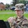 A U.S. Army soldier in Operational Camouflage Pattern (OCP) uniform is playing a trumpet outdoors. The soldier's uniform displays 'U.S. ARMY' tape and what appears to be a '132D Army Band' patch on the right shoulder. The photo is taken in a grassy outdoor setting, likely during a military band performance or rehearsal.