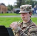 A U.S. Army soldier in Operational Camouflage Pattern (OCP) uniform is playing a trumpet outdoors. The soldier's uniform displays 'U.S. ARMY' tape and what appears to be a '132D Army Band' patch on the right shoulder. The photo is taken in a grassy outdoor setting, likely during a military band performance or rehearsal.