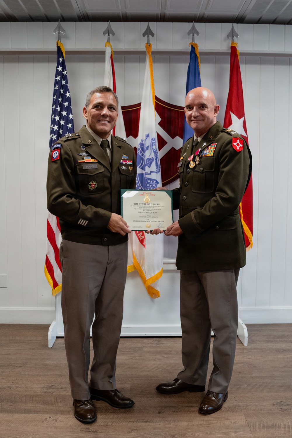 Two U.S. Army officers in dress green uniforms are jointly holding a certificate during a formal military award ceremony. The certificate appears to be 'The Florida Cross' awarded for 'Exceptionally Meritorious Service' to a Colonel. The ceremony is taking place indoors with multiple military flags displayed in the background.