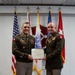 Two U.S. Army officers in dress green uniforms are jointly holding a certificate during a formal military award ceremony. The certificate appears to be 'The Florida Cross' awarded for 'Exceptionally Meritorious Service' to a Colonel. The ceremony is taking place indoors with multiple military flags displayed in the background.
