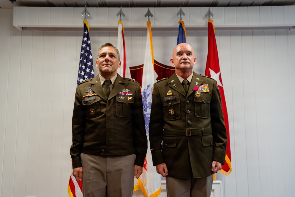 Two U.S. Army officers in dress green uniforms stand at attention before a backdrop of multiple flags including the American flag and Army unit flags. The officer on the right is wearing a medal on his chest, suggesting this is a formal military ceremony such as an award presentation or change of command. Both individuals display rank insignia and service ribbons consistent with senior officer grades.
