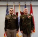 Two U.S. Army officers in dress green uniforms stand at attention before a backdrop of multiple flags including the American flag and Army unit flags. The officer on the right is wearing a medal on his chest, suggesting this is a formal military ceremony such as an award presentation or change of command. Both individuals display rank insignia and service ribbons consistent with senior officer grades.