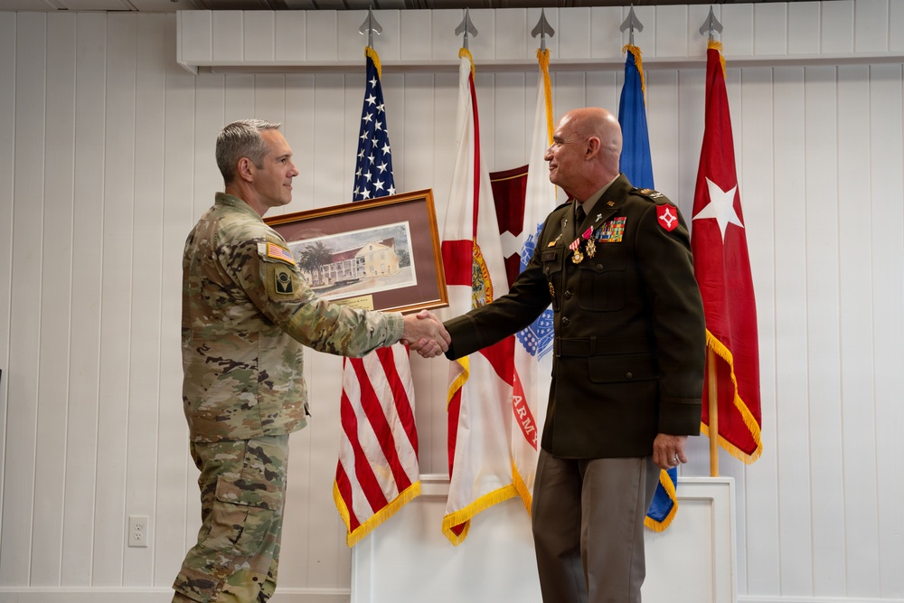 Two U.S. Army military personnel are engaged in a formal handshake ceremony, with one officer in Army Combat Uniform (ACU/OCP) presenting a framed artwork or photograph to a senior officer in Army Green Service Uniform adorned with multiple medals and ribbons. The setting appears to be an official military ceremony room with multiple flags displayed in the background. The red flag with a white star visible on the right suggests the senior officer holds a general officer rank.