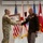 Col. Finch Concludes Distinguished Career, Leaves Lasting Legacy as Command State Chaplain Two U.S. Army military personnel are engaged in a formal handshake ceremony, with one officer in Army Combat Uniform (ACU/OCP) presenting a framed artwork or photograph to a senior officer in Army Green Service Uniform adorned with multiple medals and ribbons. The setting appears to be an official military ceremony room with multiple flags displayed in the background. The red flag with a white star visible on the right suggests the senior officer holds a general officer rank.