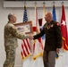 Two U.S. Army military personnel are engaged in a formal handshake ceremony, with one officer in Army Combat Uniform (ACU/OCP) presenting a framed artwork or photograph to a senior officer in Army Green Service Uniform adorned with multiple medals and ribbons. The setting appears to be an official military ceremony room with multiple flags displayed in the background. The red flag with a white star visible on the right suggests the senior officer holds a general officer rank.