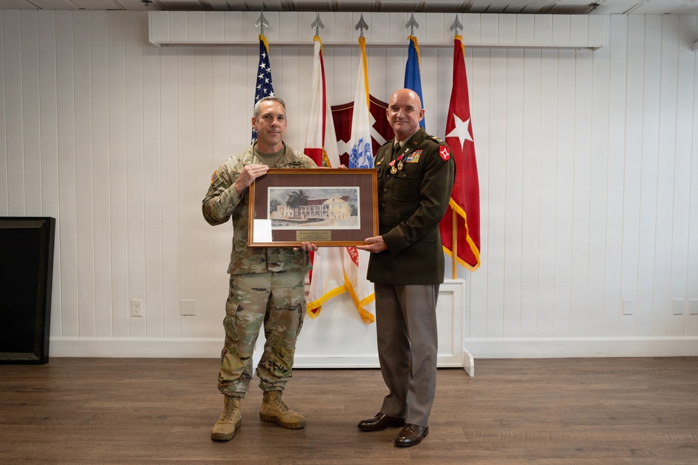 Two U.S. Army personnel are posing together while holding a framed artwork or photograph depicting a building, likely during a formal military ceremony or presentation. One individual is wearing Army Combat Uniform (ACU/OCP pattern) while the other is in Army Service Uniform (green) with multiple medals and decorations. Several military flags are displayed in the background.
