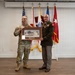 Two U.S. Army personnel are posing together while holding a framed artwork or photograph depicting a building, likely during a formal military ceremony or presentation. One individual is wearing Army Combat Uniform (ACU/OCP pattern) while the other is in Army Service Uniform (green) with multiple medals and decorations. Several military flags are displayed in the background.