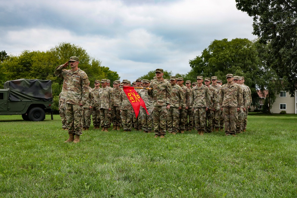 A military unit is assembled in formation on a grass field, with one soldier in the foreground rendering a salute. A guidon bearer holds a red pennant flag with yellow markings in the center of the formation. The setting appears to be a formal military ceremony or parade on a post or installation.