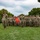 A military unit is assembled in formation on a grass field, with one soldier in the foreground rendering a salute. A guidon bearer holds a red pennant flag with yellow markings in the center of the formation. The setting appears to be a formal military ceremony or parade on a post or installation.