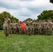 A military unit is assembled in formation on a grass field, with one soldier in the foreground rendering a salute. A guidon bearer holds a red pennant flag with yellow markings in the center of the formation. The setting appears to be a formal military ceremony or parade on a post or installation.