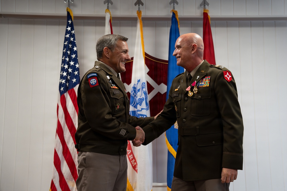 Two U.S. Army officers in dress green uniforms are shaking hands during what appears to be a formal military ceremony. The officer on the left wears an 82nd Airborne Division patch, while the officer on the right displays multiple medals and ribbons along with a chaplain's cross insignia. Several military and U.S. flags are displayed in the background.