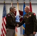 Two U.S. Army officers in dress green uniforms are shaking hands during what appears to be a formal military ceremony. The officer on the left wears an 82nd Airborne Division patch, while the officer on the right displays multiple medals and ribbons along with a chaplain's cross insignia. Several military and U.S. flags are displayed in the background.