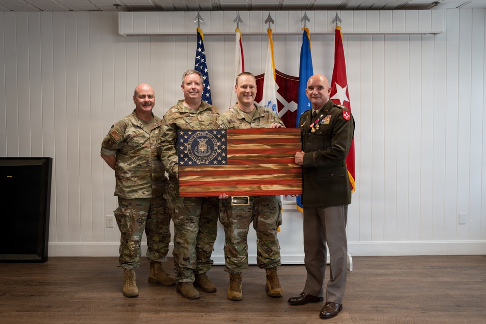 Four military personnel pose for a formal photograph in what appears to be an indoor ceremony room. Two of the individuals are holding a wooden American flag plaque bearing a United States Air Force seal. The group includes three individuals in Army Combat Uniform (ACU/OCP) and one in a formal dress uniform consistent with the United States Marine Corps.