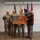 Col. Finch Concludes Distinguished Career, Leaves Lasting Legacy as Command State Chaplain Four military personnel pose for a formal photograph in what appears to be an indoor ceremony room. Two of the individuals are holding a wooden American flag plaque bearing a United States Air Force seal. The group includes three individuals in Army Combat Uniform (ACU/OCP) and one in a formal dress uniform consistent with the United States Marine Corps.