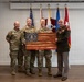 Four military personnel pose for a formal photograph in what appears to be an indoor ceremony room. Two of the individuals are holding a wooden American flag plaque bearing a United States Air Force seal. The group includes three individuals in Army Combat Uniform (ACU/OCP) and one in a formal dress uniform consistent with the United States Marine Corps.