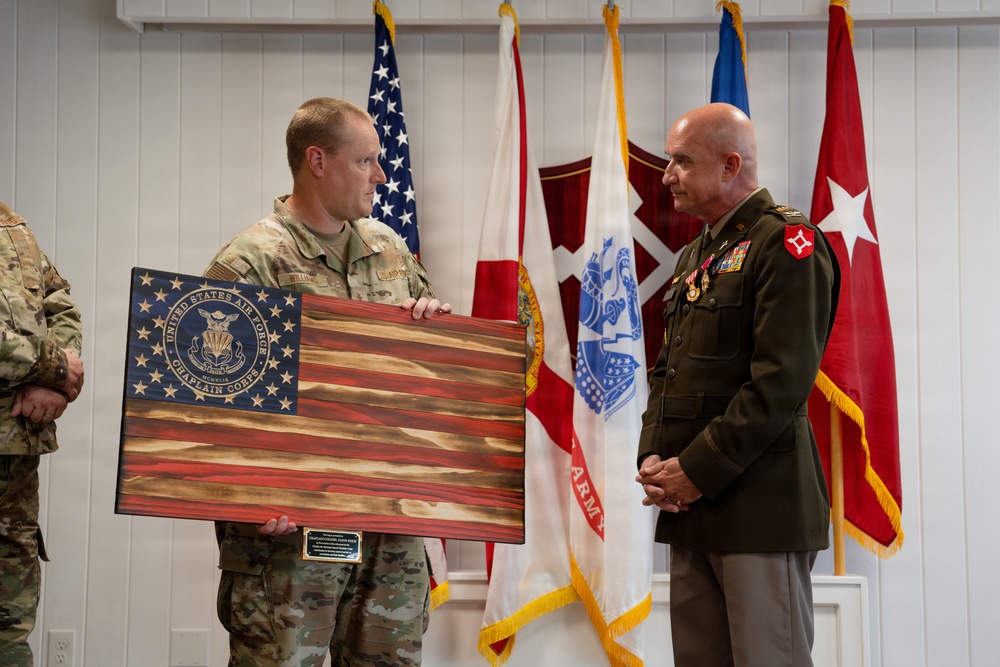 A military ceremony in which a soldier in Army Combat Uniform presents a wooden American flag plaque bearing the United States Air Force Chaplain Corps insignia to a senior officer in Marine Corps dress greens. The recipient displays multiple medals and ribbons along with general officer rank insignia. Several military flags are displayed in the background, indicating a formal military setting.