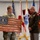 Col. Finch Concludes Distinguished Career, Leaves Lasting Legacy as Command State Chaplain A military ceremony in which a soldier in Army Combat Uniform presents a wooden American flag plaque bearing the United States Air Force Chaplain Corps insignia to a senior officer in Marine Corps dress greens. The recipient displays multiple medals and ribbons along with general officer rank insignia. Several military flags are displayed in the background, indicating a formal military setting.