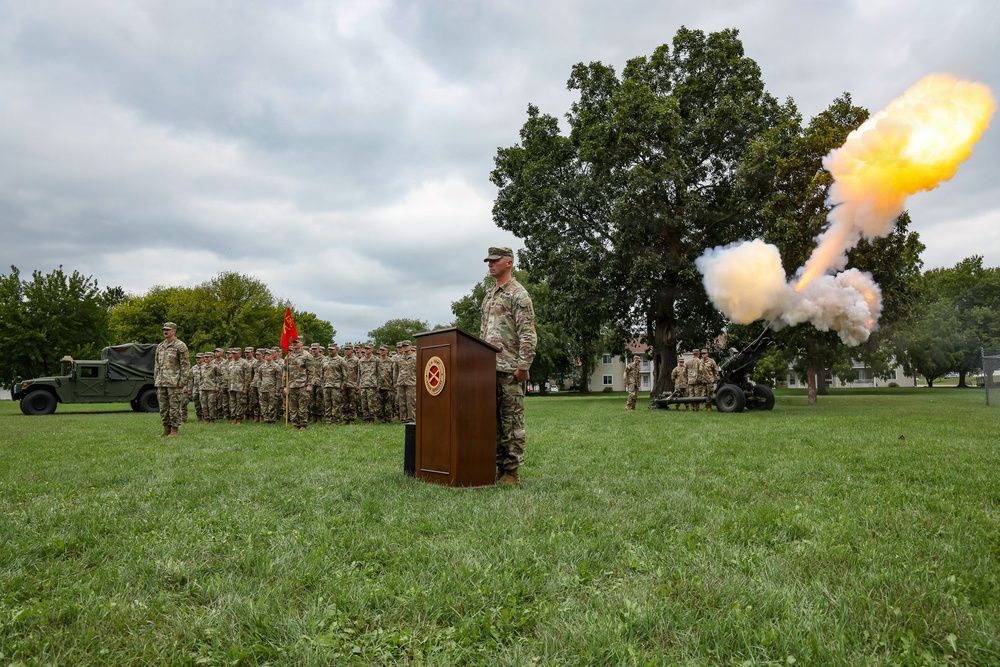 A formal U.S. Army military ceremony is taking place on a grassy parade field, featuring a cannon salute firing in progress. A soldier stands at a wooden podium bearing a military insignia emblem, while a formation of soldiers in Army Combat Uniforms stands at attention in the background. A HMMWV (Humvee) and an artillery piece (howitzer) are also visible, consistent with a change of command or similar ceremonial event.