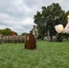 A formal U.S. Army military ceremony is taking place on a grassy parade field, featuring a cannon salute firing in progress. A soldier stands at a wooden podium bearing a military insignia emblem, while a formation of soldiers in Army Combat Uniforms stands at attention in the background. A HMMWV (Humvee) and an artillery piece (howitzer) are also visible, consistent with a change of command or similar ceremonial event.